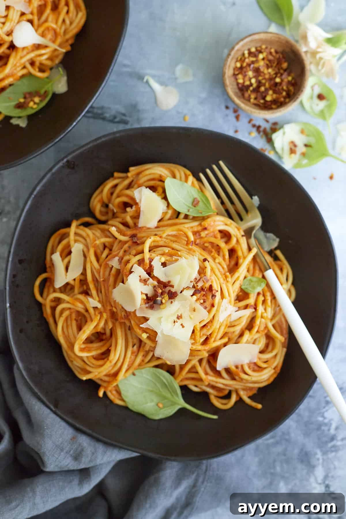 A serving of arrabiata sauce spaghetti on a black plate topped with fresh Parmesan cheese, basil leaves, and red pepper flakes with a fork on the side.