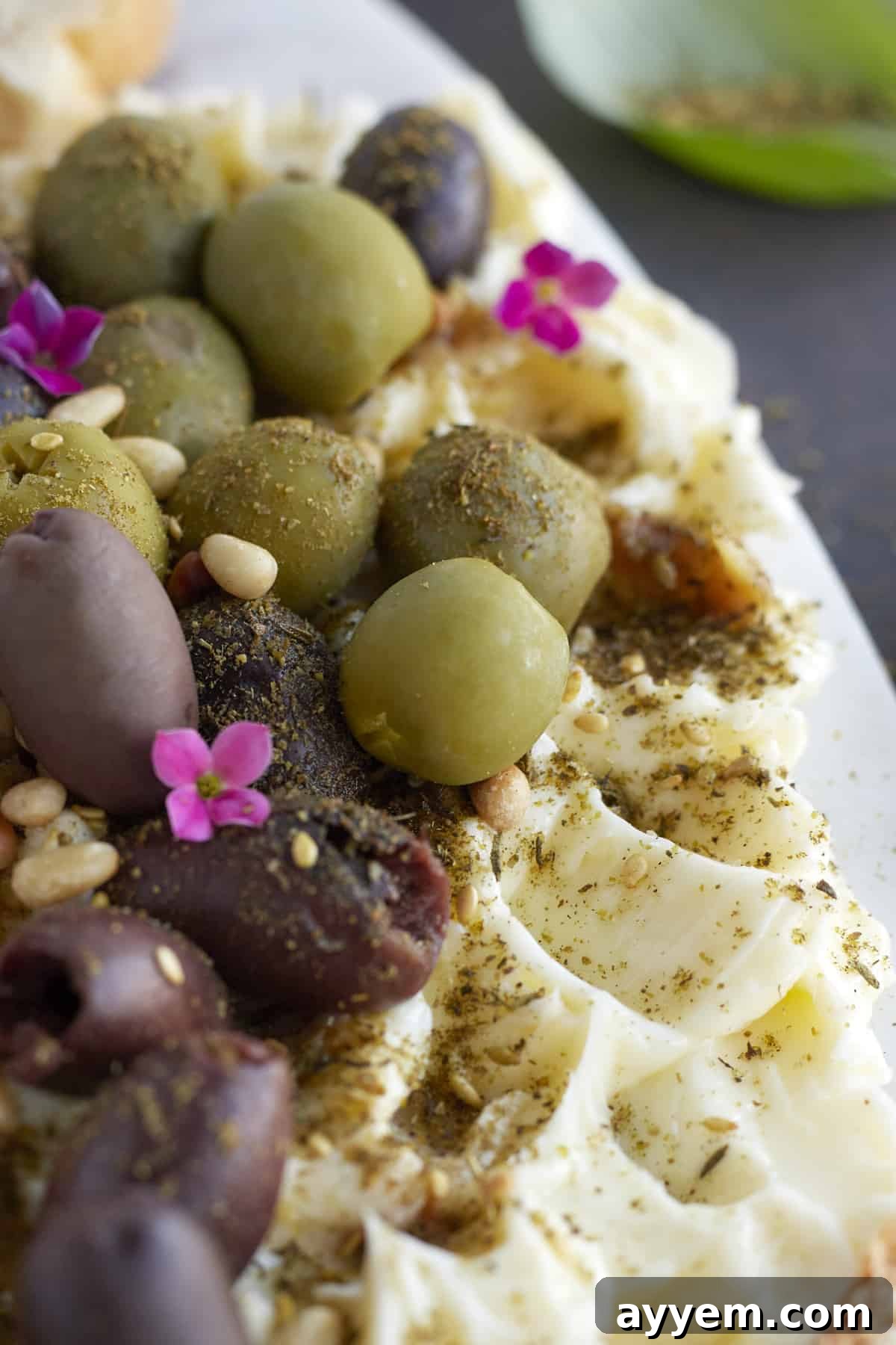 Close-up image of a butter board featuring creamy butter, an assortment of green and black olives, toasted pine nuts, roasted garlic cloves, and a sprinkle of seasonings.