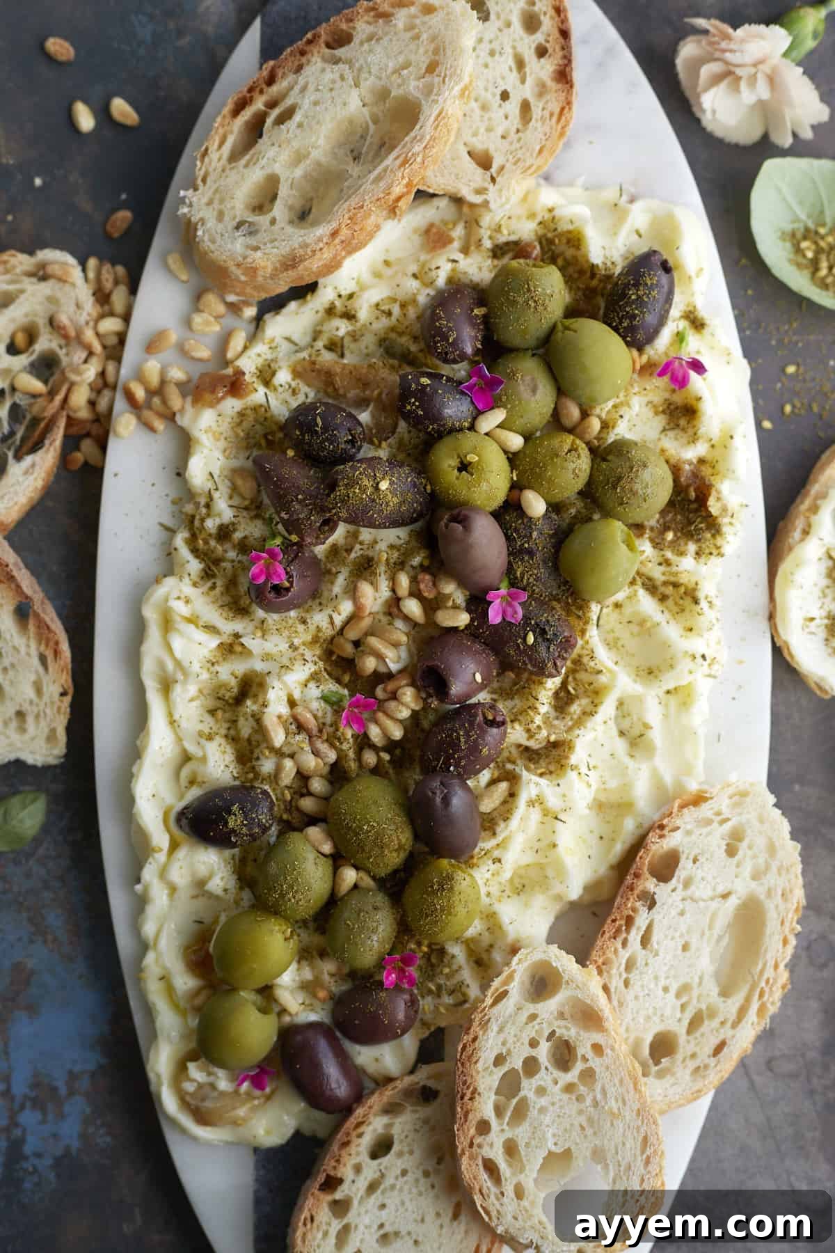 Overhead image of a butter board topped with green and black olives, pine nuts, and seasonings with crostini around the sides, a perfect savory appetizer.