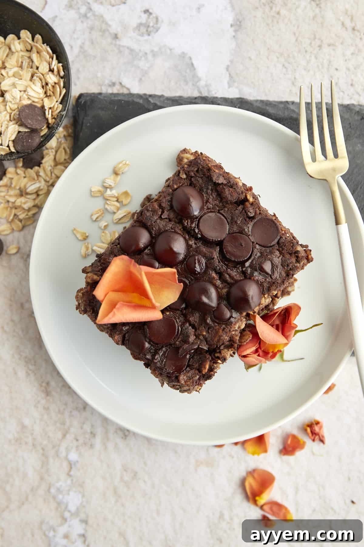 An inviting overhead view displays a single slice of chocolate baked oats, elegantly arranged on a pristine white plate and adorned with delicate orange flower petals. A fork rests gently alongside, beckoning a taste of this wholesome, sweet breakfast.