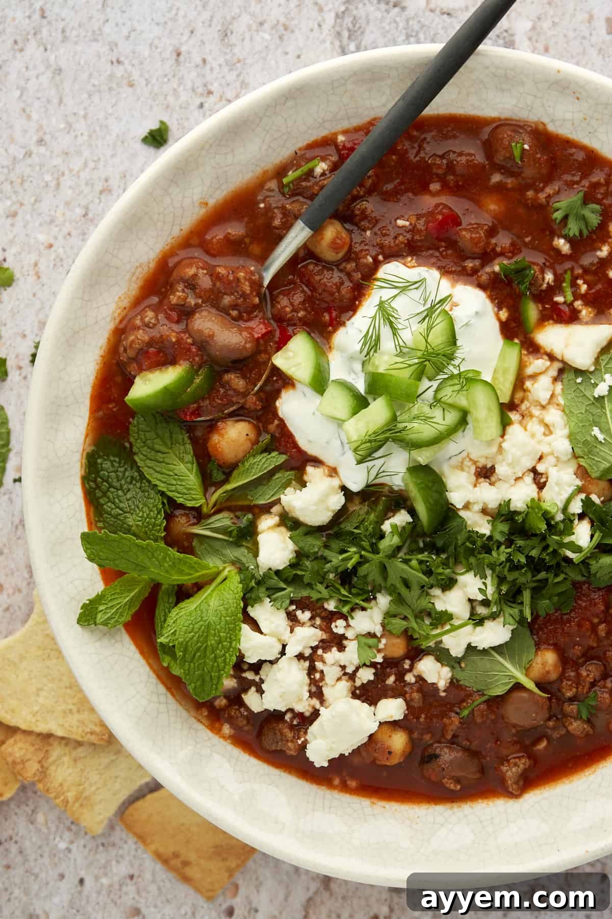 A beautifully presented white bowl of Middle Eastern beef chili, garnished with a swirl of yogurt sauce, crumbled feta cheese, and fresh green herbs.