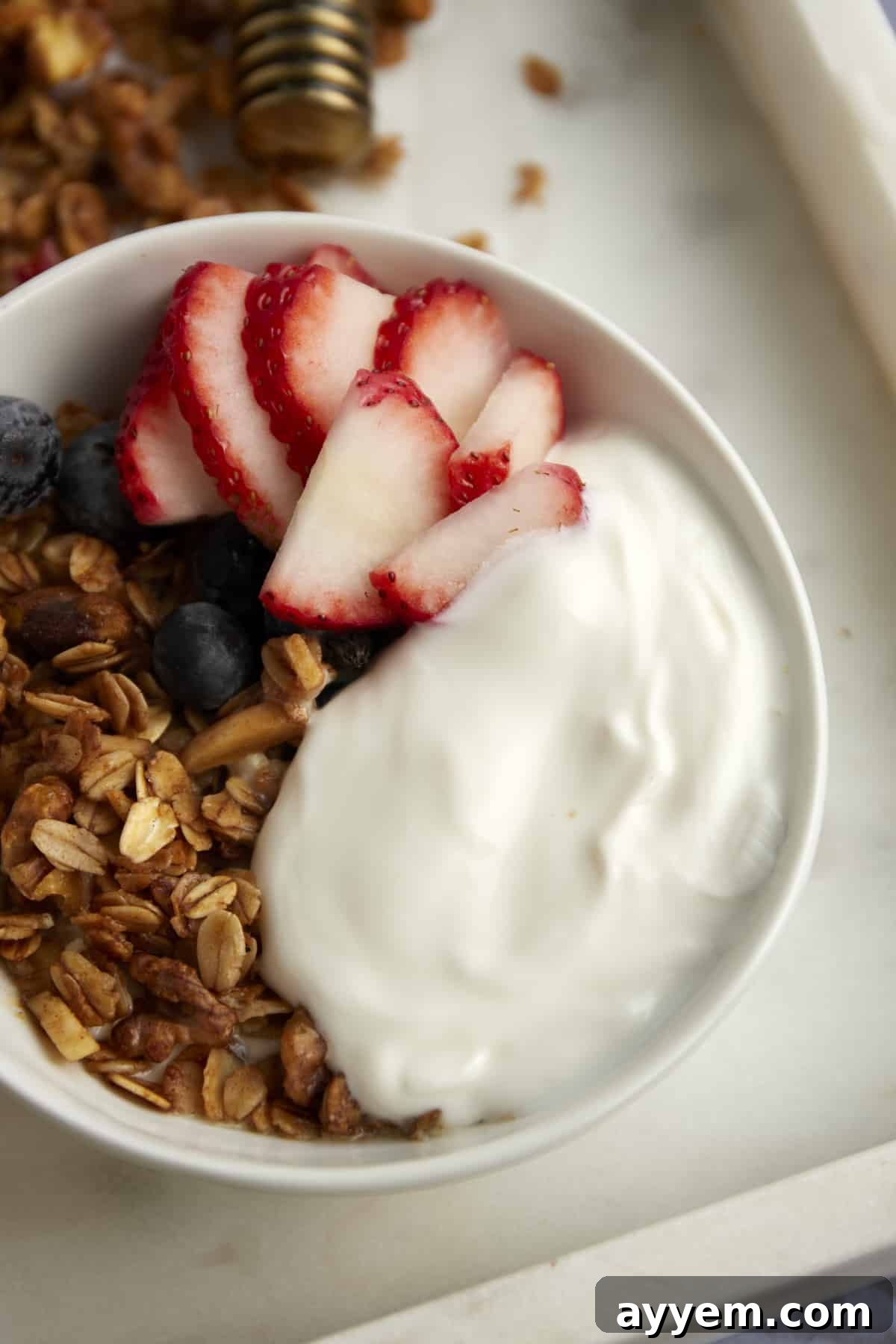 A white bowl showcasing Baklava Granola generously topped over creamy yogurt, adorned with fresh strawberries and blueberries.