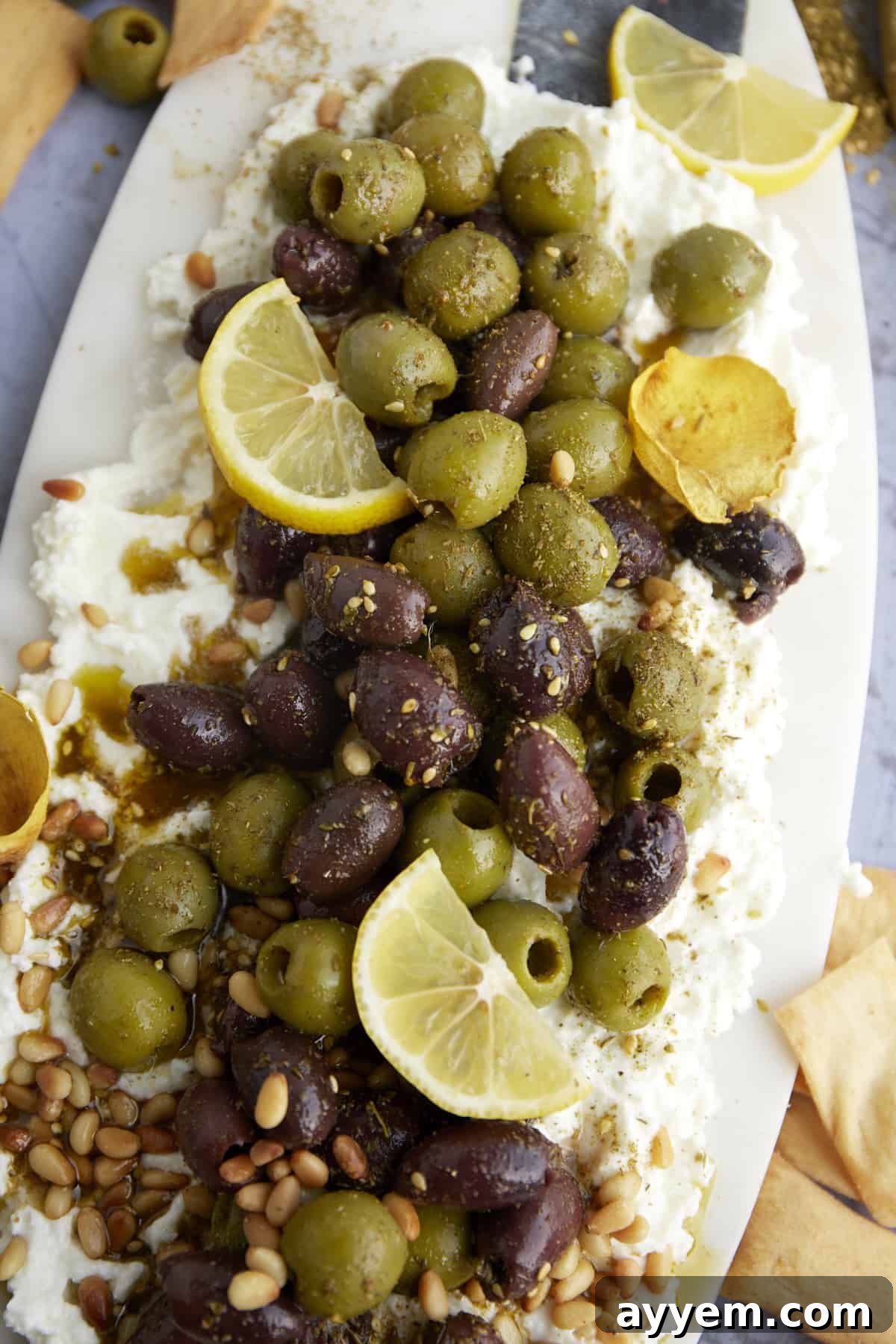 A close-up view of a beautifully arranged feta board, showcasing a colorful mix of marinated Kalamata and green olives, garnished with lemon wedges and aromatic herbs, ready for dipping.