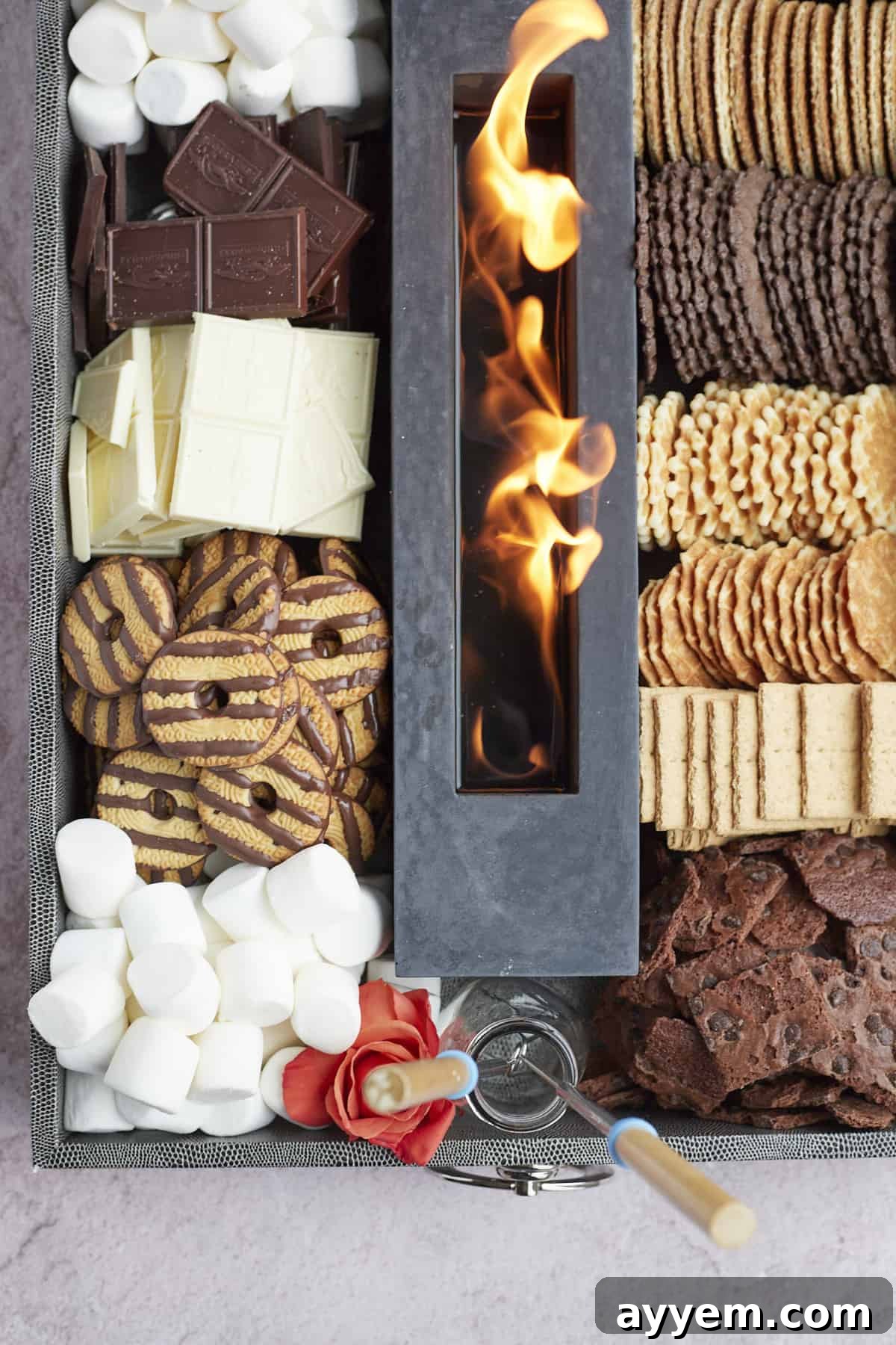 An overhead shot of a large, lavish indoor s'mores board, featuring a central portable fire pit surrounded by a variety of cookies, marshmallows, graham crackers, different chocolates, and fresh fruit, beautifully arranged for a crowd.