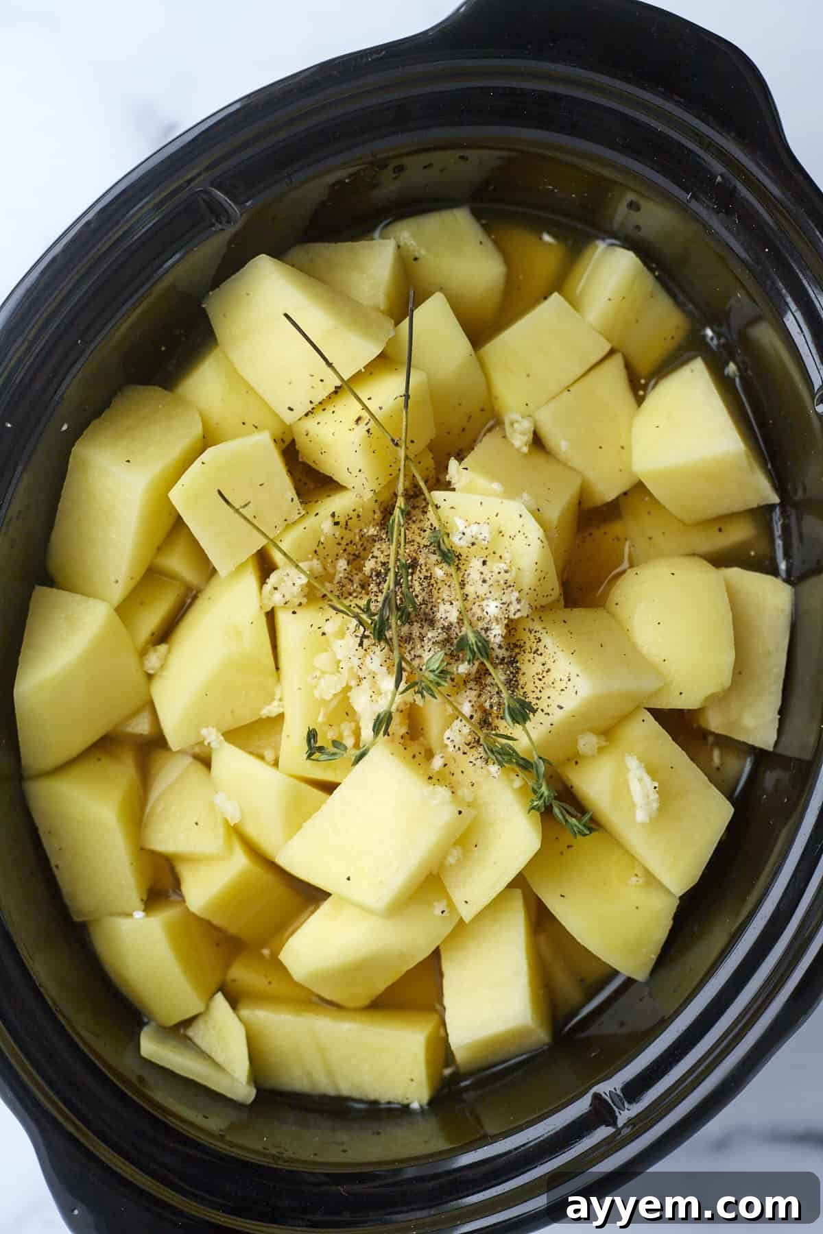 Cubed potatoes, aromatic garlic, and fresh herbs submerged in vegetable broth within a slow cooker, ready for cooking.