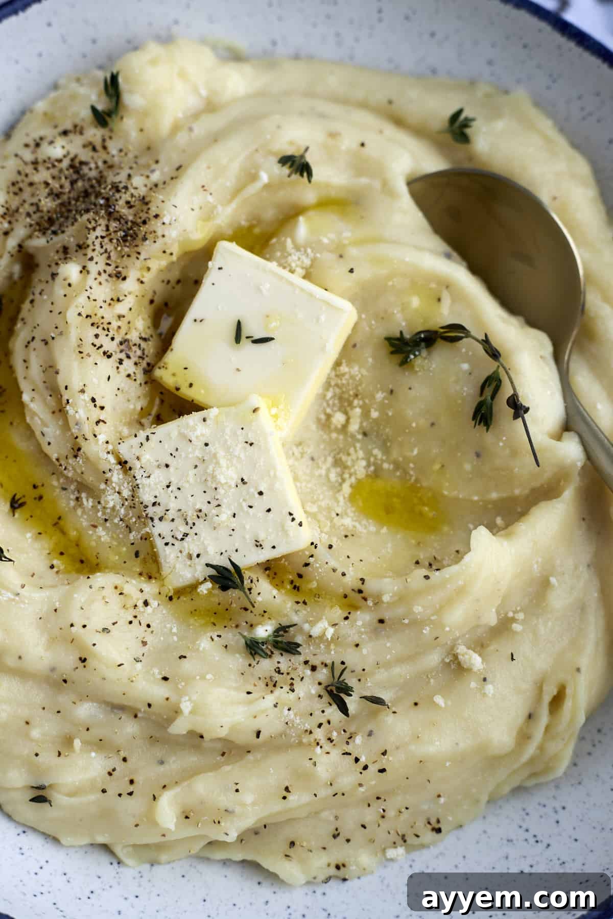 A close-up view of creamy slow cooker mashed potatoes in a bowl, generously topped with two pads of butter, grated Parmesan cheese, and fresh cracked black pepper.