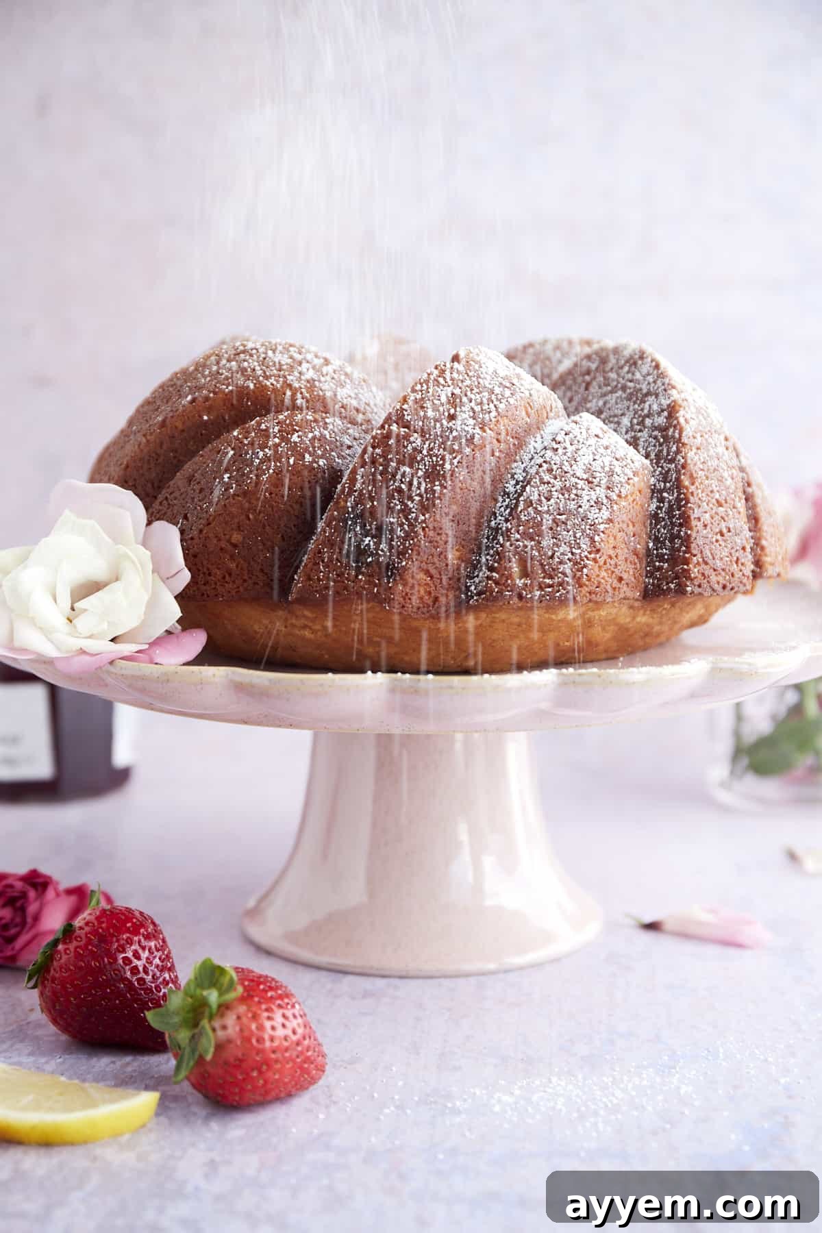 A beautifully baked Strawberry Lemon Bundt Cake on a cake stand, being dusted with powdered sugar, ready to be served.