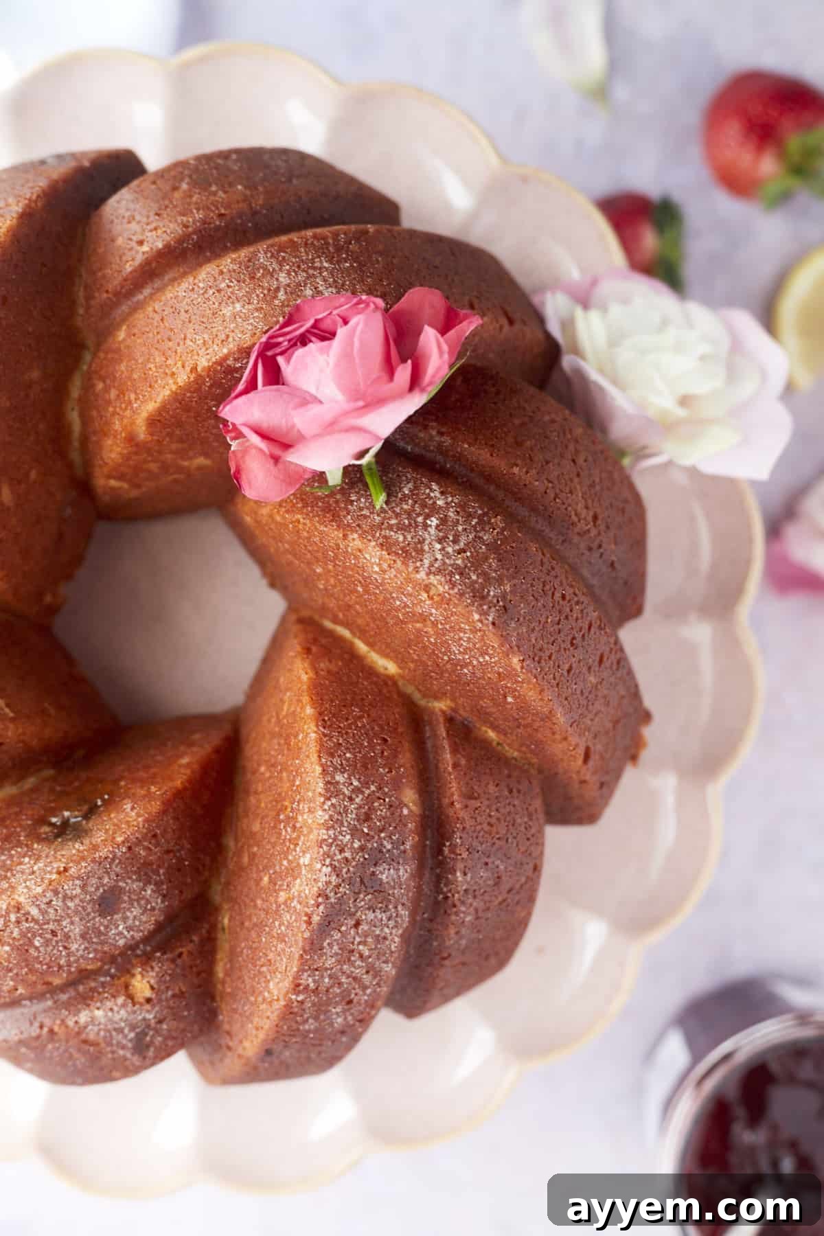 An overhead view of a plain, freshly baked strawberry lemon bundt cake on a white cake stand, adorned with a single pink rose.