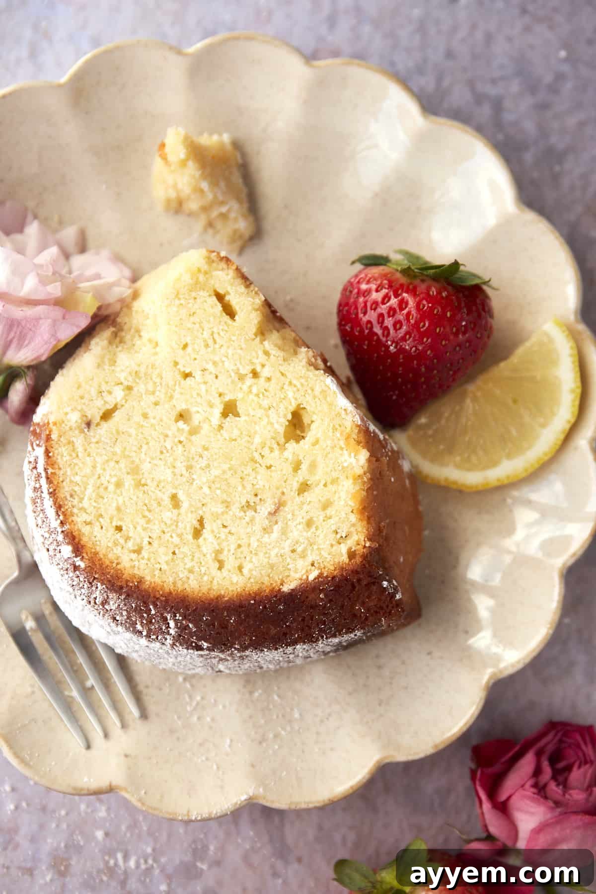 An overhead view of a slice of bundt cake, topped with powdered sugar, on a white plate with a fork, a fresh strawberry, and a lemon slice.
