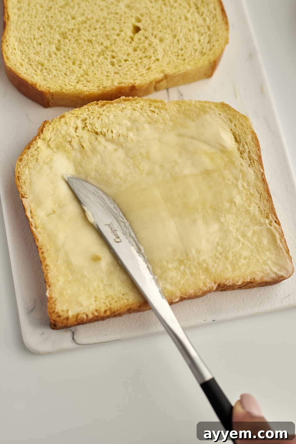 A close-up of two pieces of bread, with a hand gently buttering one slice, showcasing the preparation of this cinnamon French toast.