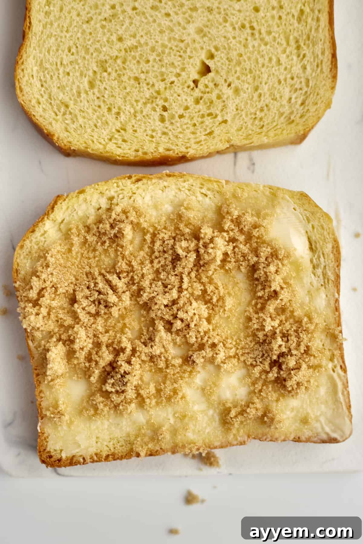 Two golden slices of brioche bread, one perfectly buttered and generously topped with brown sugar, laid out in preparation for the baked cinnamon French toast casserole.