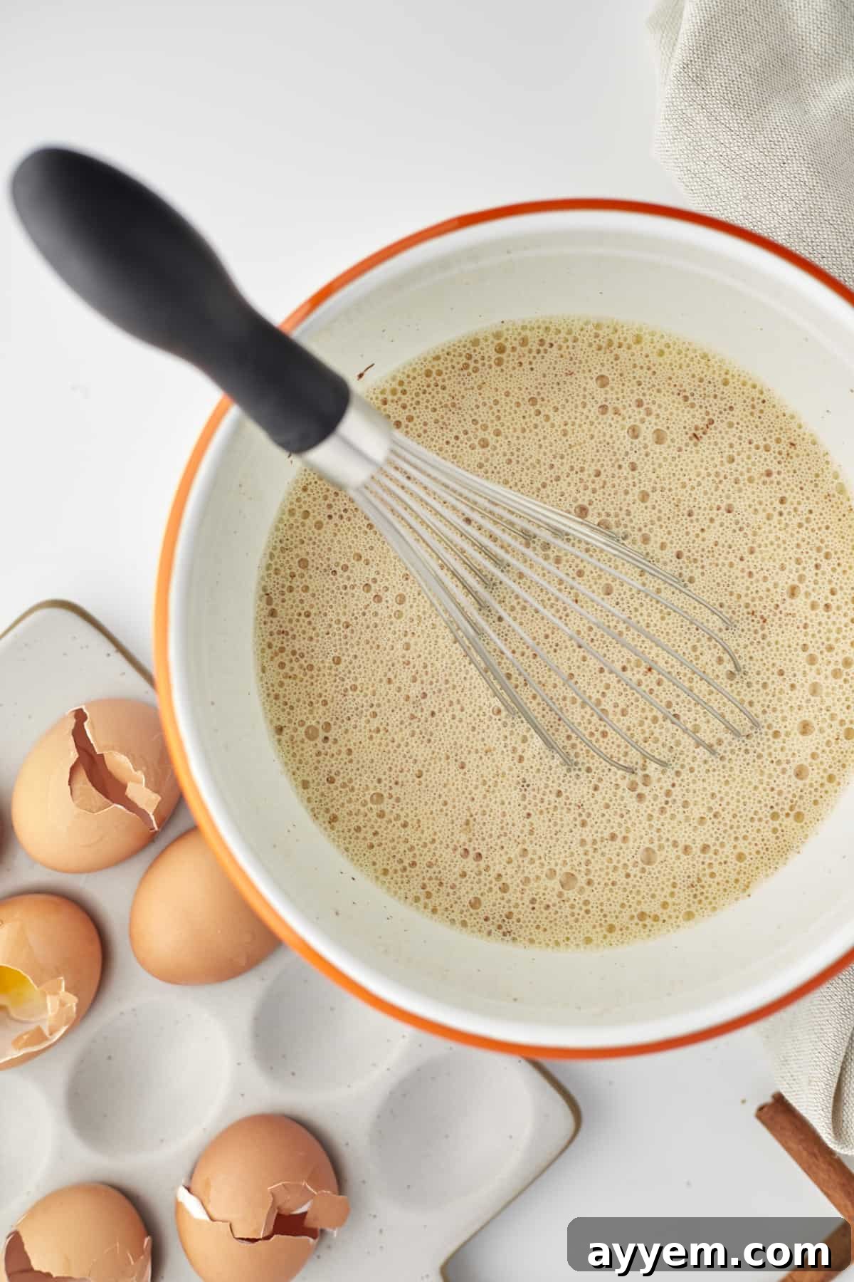 An overhead shot showcasing a bowl filled with a perfectly whisked egg mixture, surrounded by fresh eggs, ready for preparing a delicious baked cinnamon French toast recipe.
