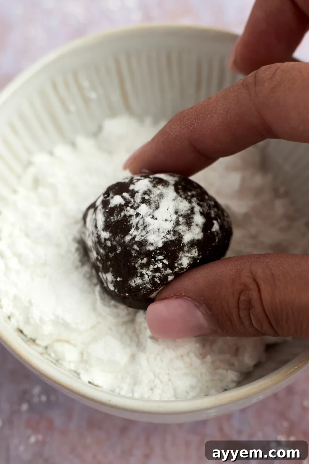 Two fingers gently rolling a ball of chocolate crinkle cookie dough in a bowl of powdered sugar, demonstrating the preparation step.