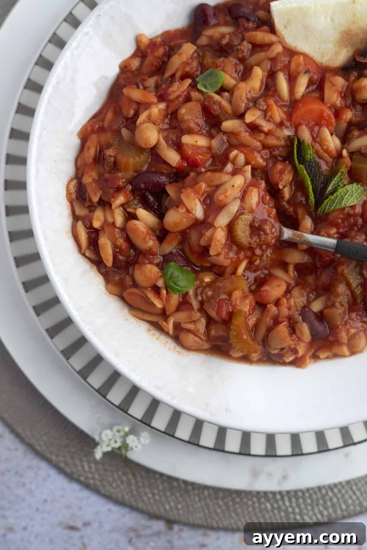 Overhead shot of a full bowl of Pasta e Fagioli, garnished with fresh herbs, with a spoon and bread slices nearby.