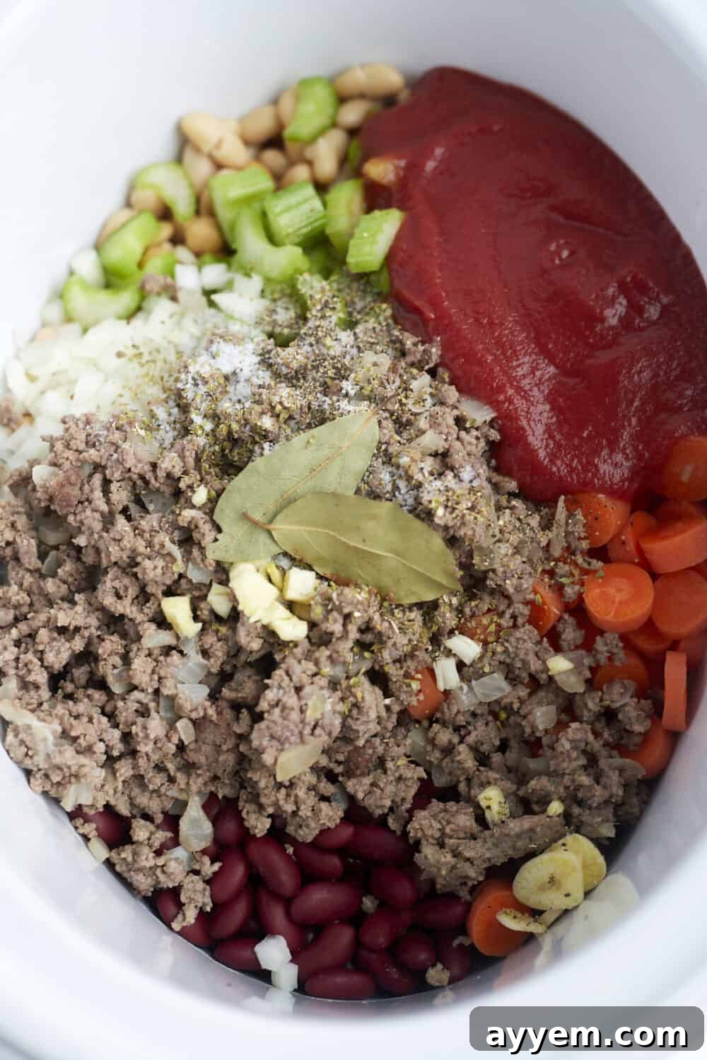 A slow cooker filled with uncooked ingredients for pasta e fagioli, including ground beef, diced vegetables, various beans, tomato sauce, and seasonings, ready to be cooked.