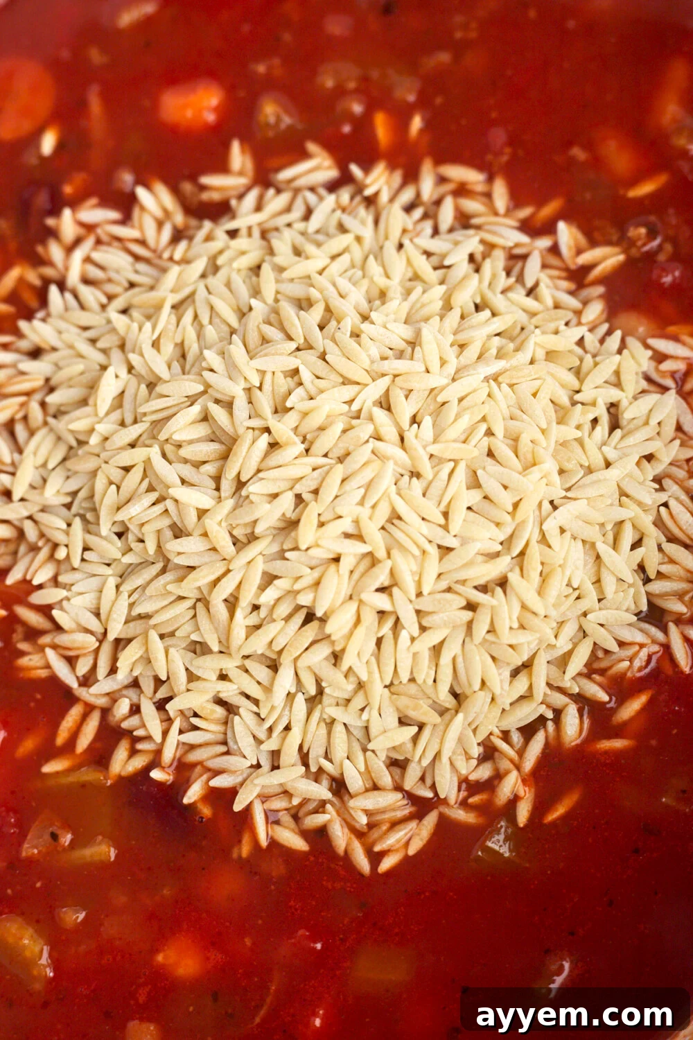 Uncooked orzo pasta being sprinkled over a rich, red pasta sauce in a slow cooker, indicating the final stage of ingredient addition.