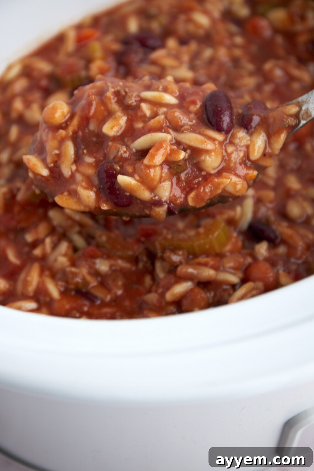 A spoon scooping a generous portion of thick pasta and beans soup directly from a slow cooker, emphasizing its hearty consistency.