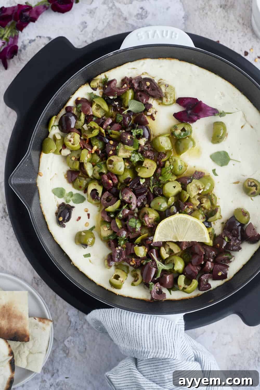 A vibrant warm feta dip generously topped with glistening marinated olives and fresh herbs, ready for serving.