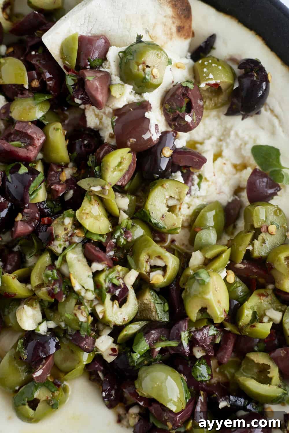 Close-up of crusty bread being dipped into a luscious warm feta dip, topped with colorful marinated olives.