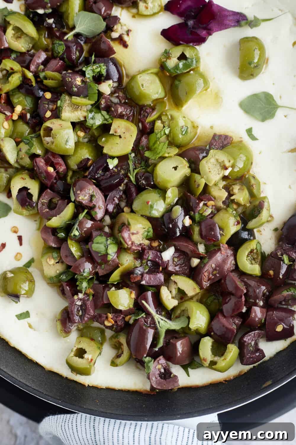 A top-down shot of a rustic warm feta dip in a skillet, adorned with colorful marinated olives, fresh herbs, and a drizzle of olive oil.