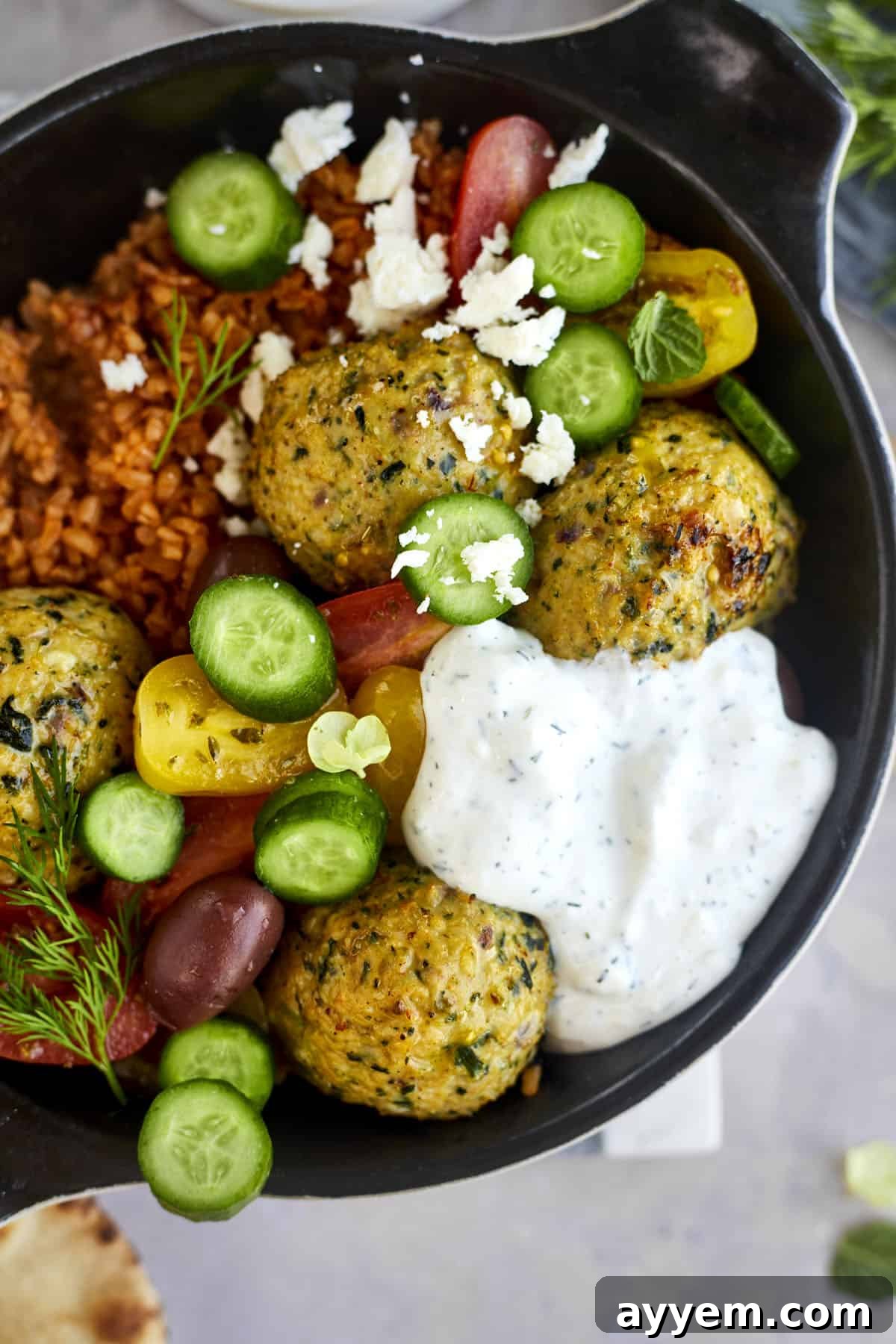 Greek meatballs in a skillet with rice, cucumbers, tomatoes, tzatziki, and feta