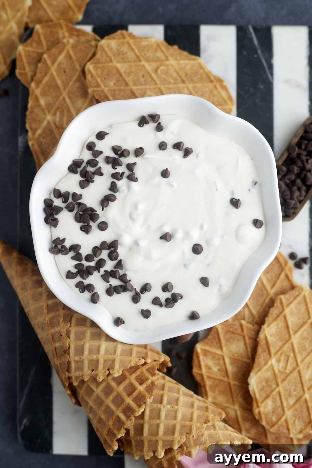A close-up of a bowl of homemade cannoli dip, generously topped with mini chocolate chips, set against a backdrop of waffle cones and cookies, inviting a delicious scoop.
