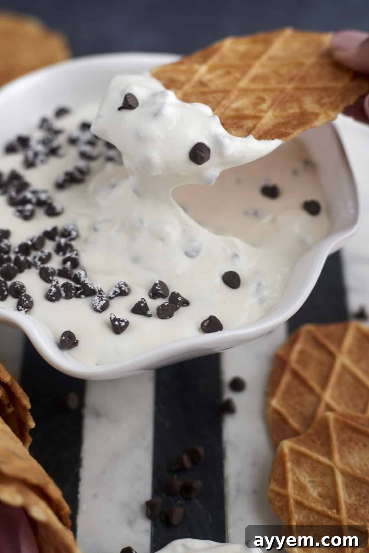 A hand dipping a crispy waffle cone piece into a rich, creamy bowl of cannoli dip, showing the delicious texture and chocolate chip filling.