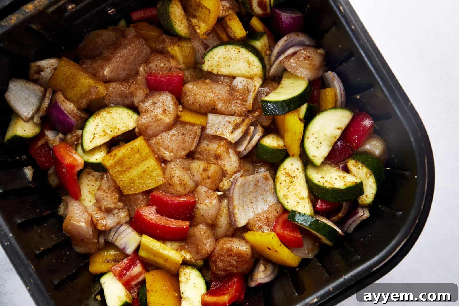 Raw chicken breast pieces and colorful chopped vegetables arranged in an air fryer basket, ready for cooking.