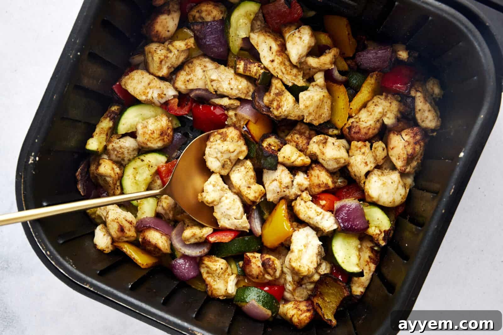 A spoon scooping up perfectly cooked air fryer chicken breast and colorful vegetables from an air fryer basket.