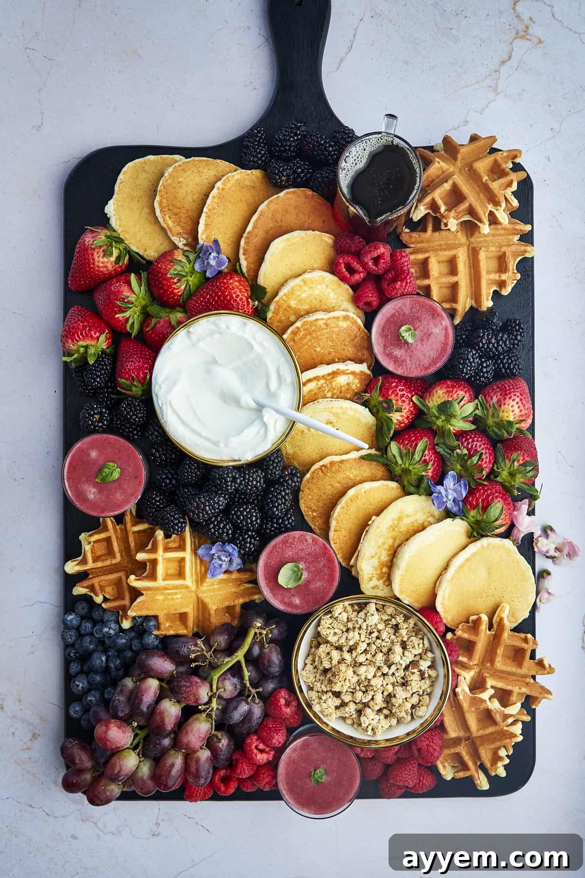 A vibrant breakfast charcuterie board piled high with pancakes, waffles, fresh berries, bacon, and various toppings, perfect for a festive brunch.
