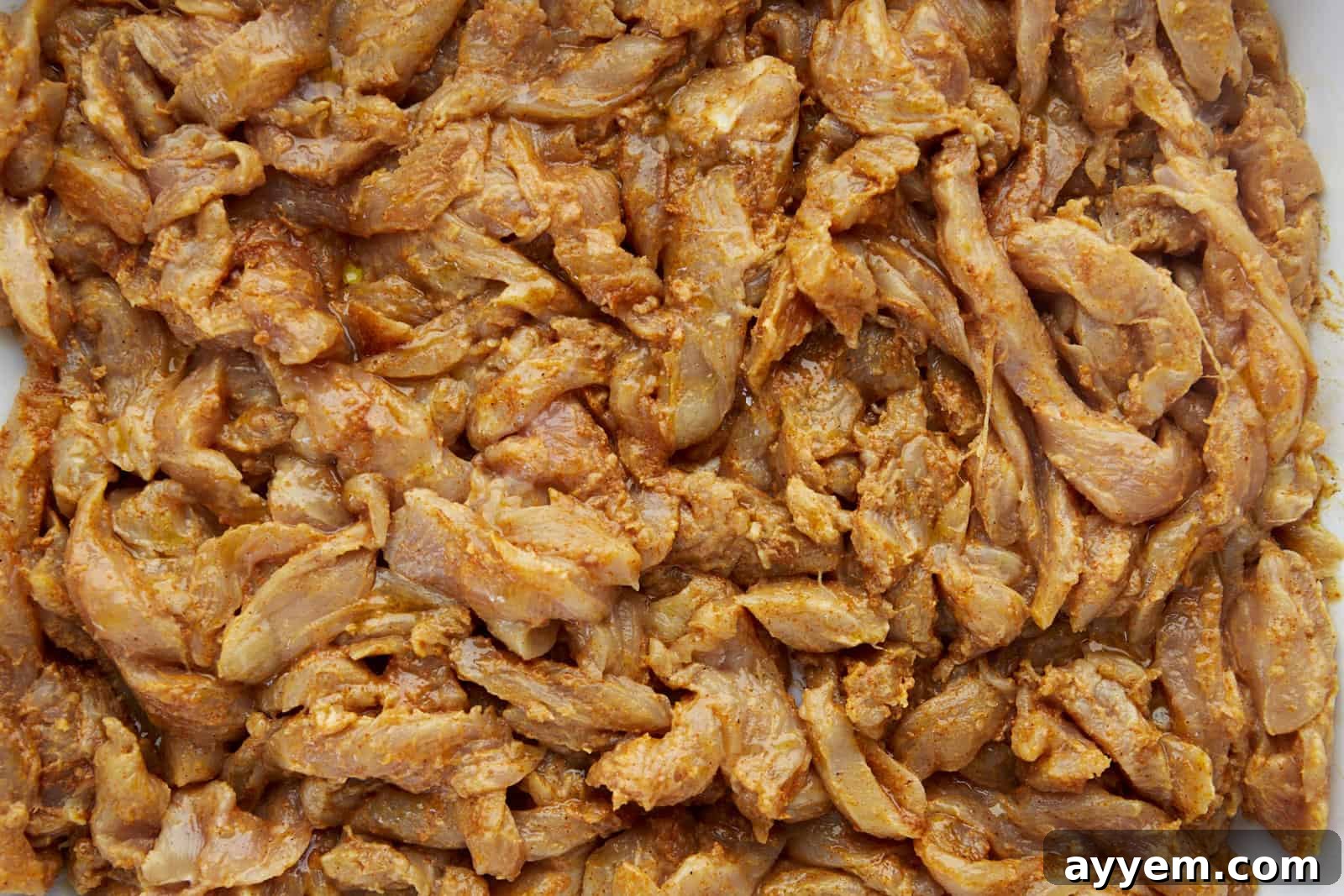 Raw chicken breast slices coated generously in a vibrant, creamy shawarma marinade, ready for baking. The rich orange hue of the spices promises deep flavor.