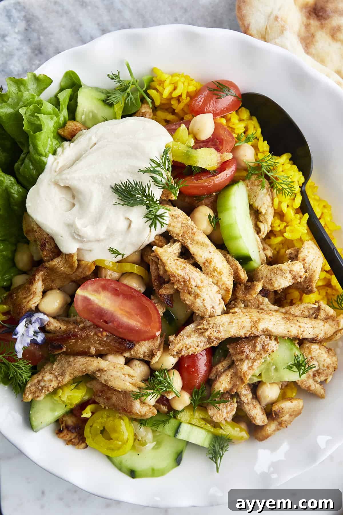 A beautifully plated bowl of shawarma chicken, nestled beside fluffy yellow rice, crisp cucumbers, ripe tomatoes, chickpeas, fresh lettuce, and a swirl of creamy hummus, embodying a complete and satisfying Middle Eastern meal.