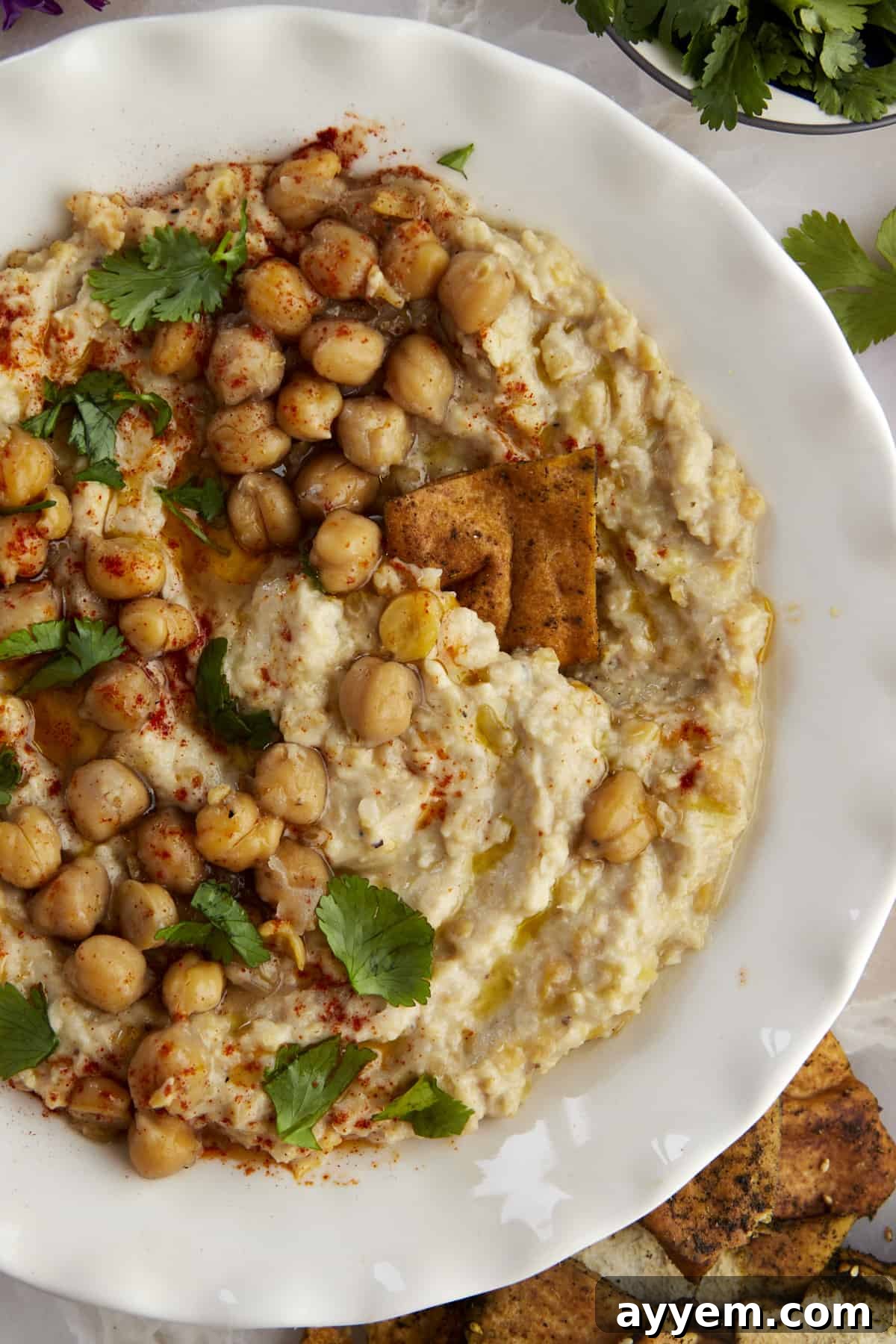 A hand dipping a crispy pita chip into a bowl of homemade chunky garlic hummus (Masabaha), garnished with whole chickpeas and olive oil.