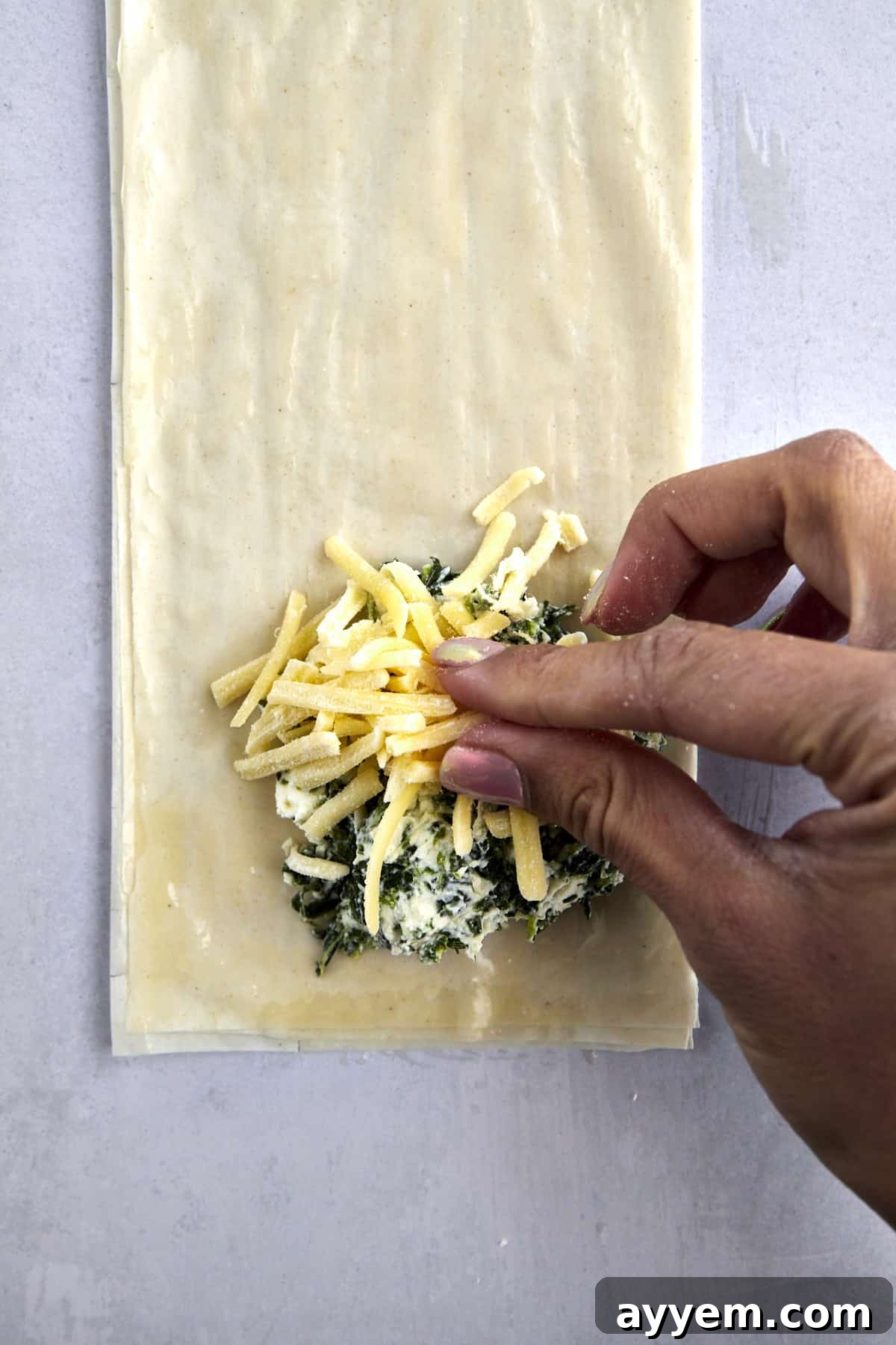 A hand sprinkling cheese over a spinach spanakopita filling over layered phyllo dough. 