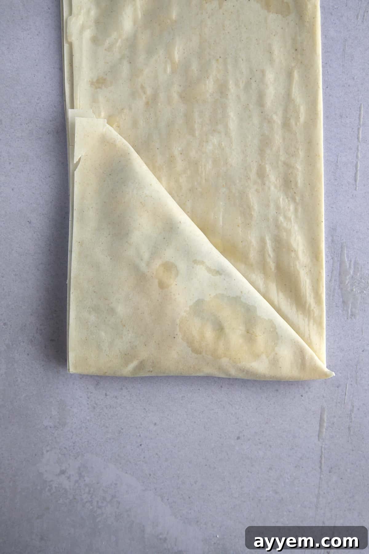 Raw layered phyllo dough sheets stuffed with spanakopita filling being folded into a triangle. 