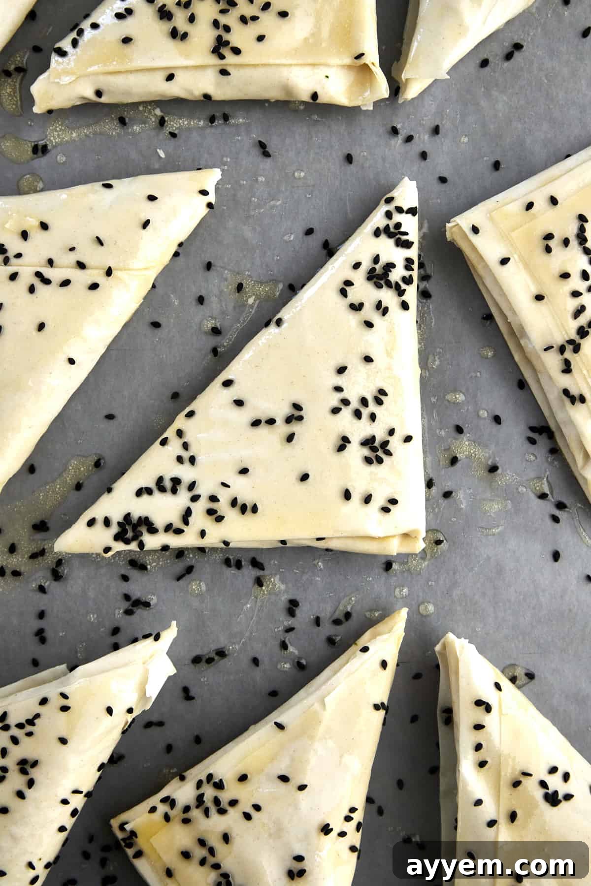 Raw spanakopita triangles on a baking sheet topped with butter and black sesame seeds. 