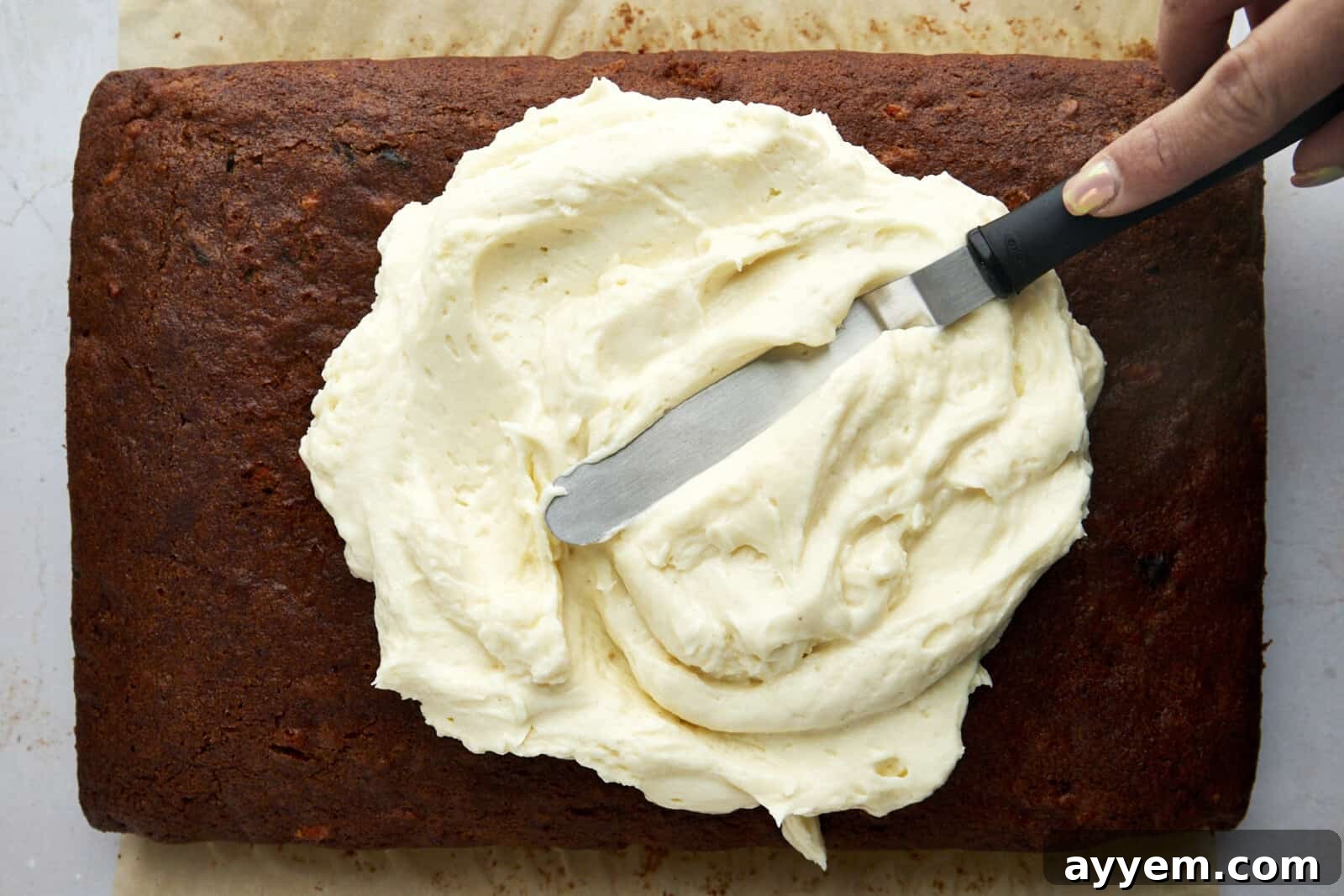 Cream cheese frosting being expertly spread over a cooled carrot cake using an offset spatula.
