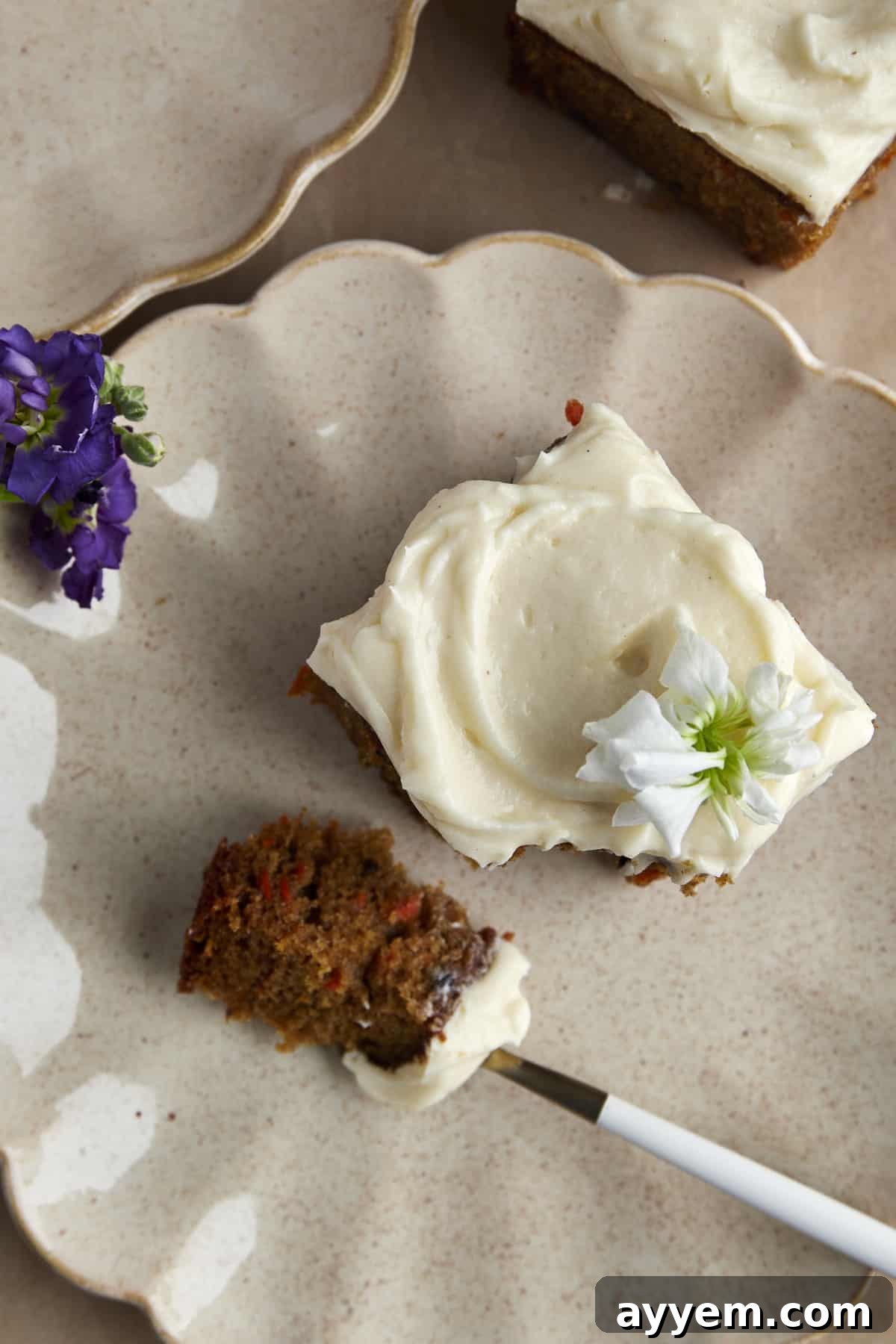 A square piece of carrot cake topped with cream cheese frosting on a white plate, with a fork holding a perfect bite.