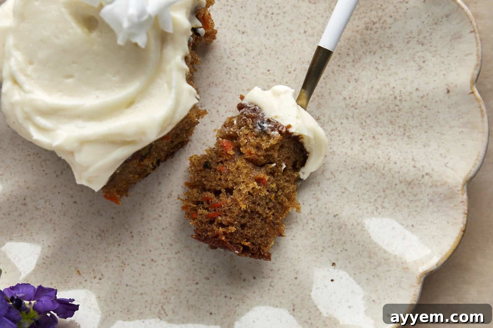A forkful of moist carrot cake with vanilla bean cream cheese frosting on a plate, next to the remaining slice.