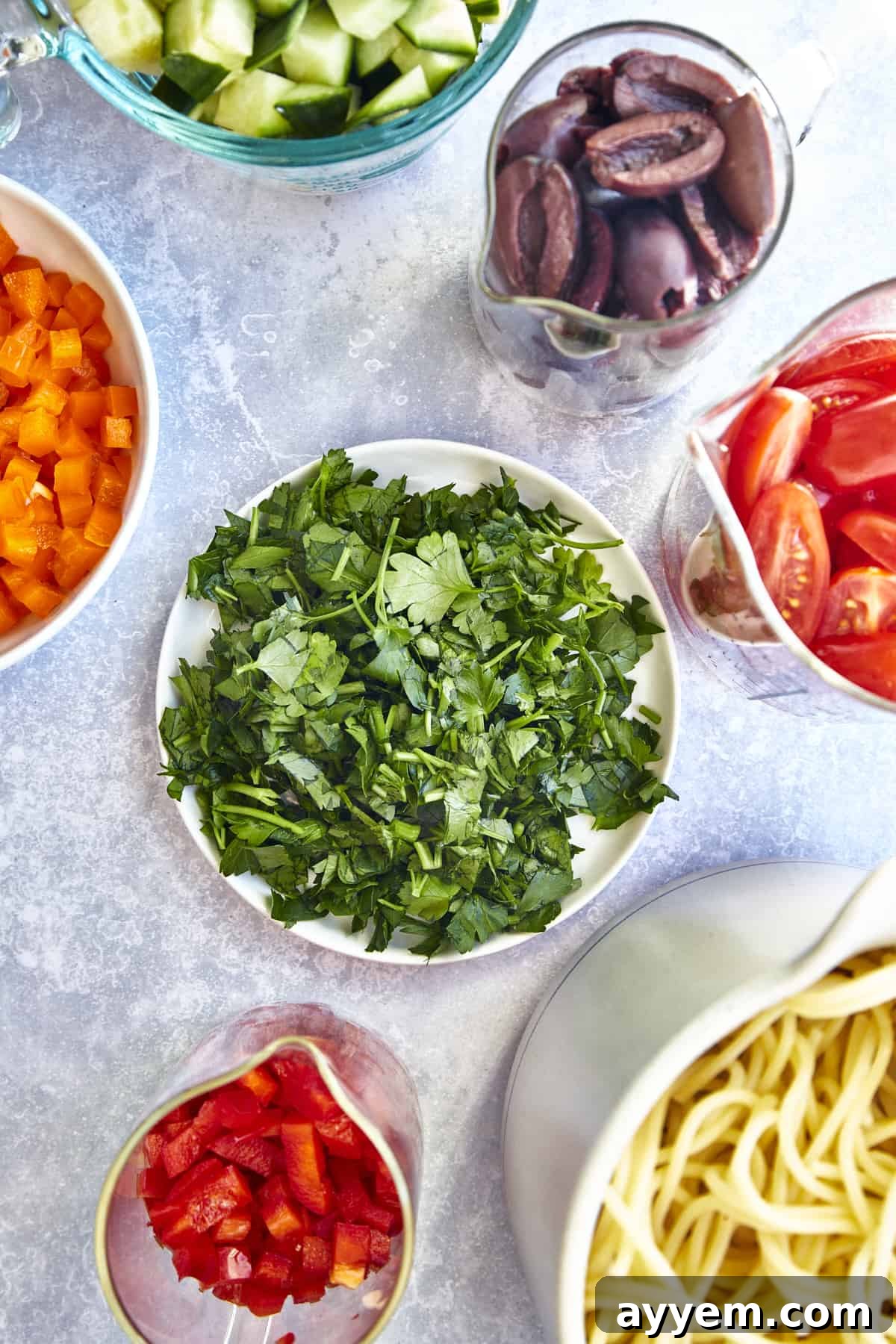 Bowls of kalamata olives, halved tomatoes, chopped red and orange bell peppers, cooked spaghetti noodles, and fresh parsley. 