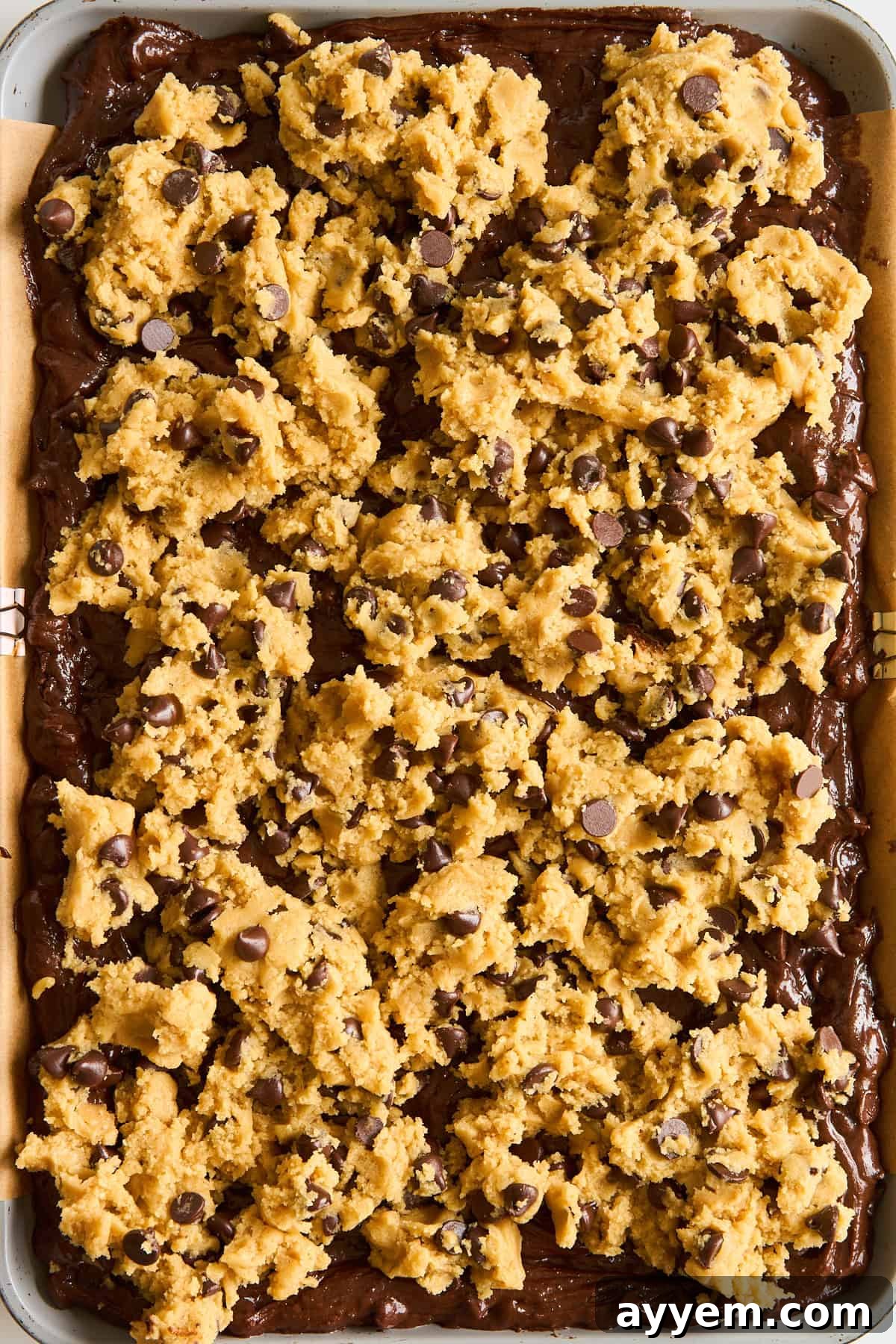 A baking tray filled with brownie batter topped with uneven scoops of chocolate chip cookie dough, ready to be baked.