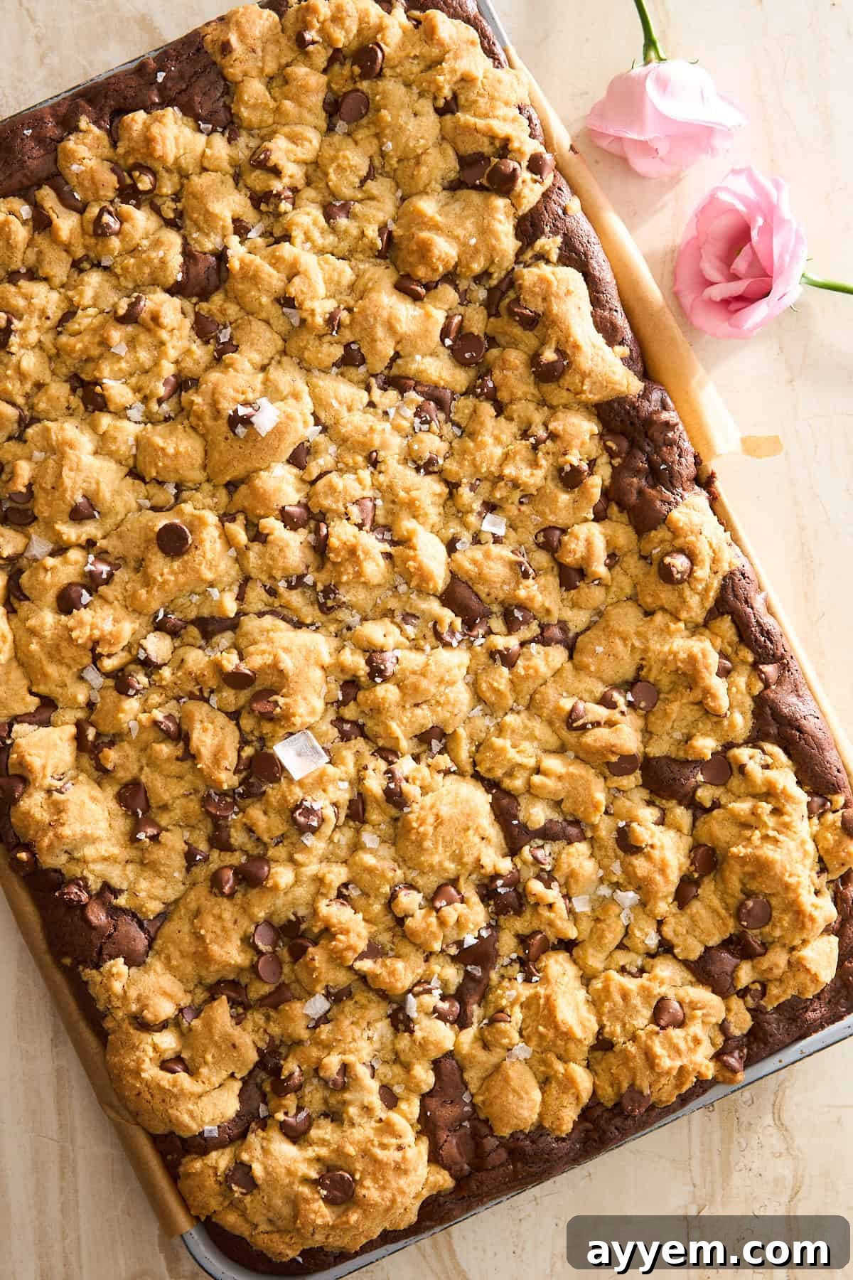 A large tray of chocolate chip cookie bars with a golden, crumbly topping and chocolate chips, set on parchment paper with two pink flowers nearby on a light surface.
