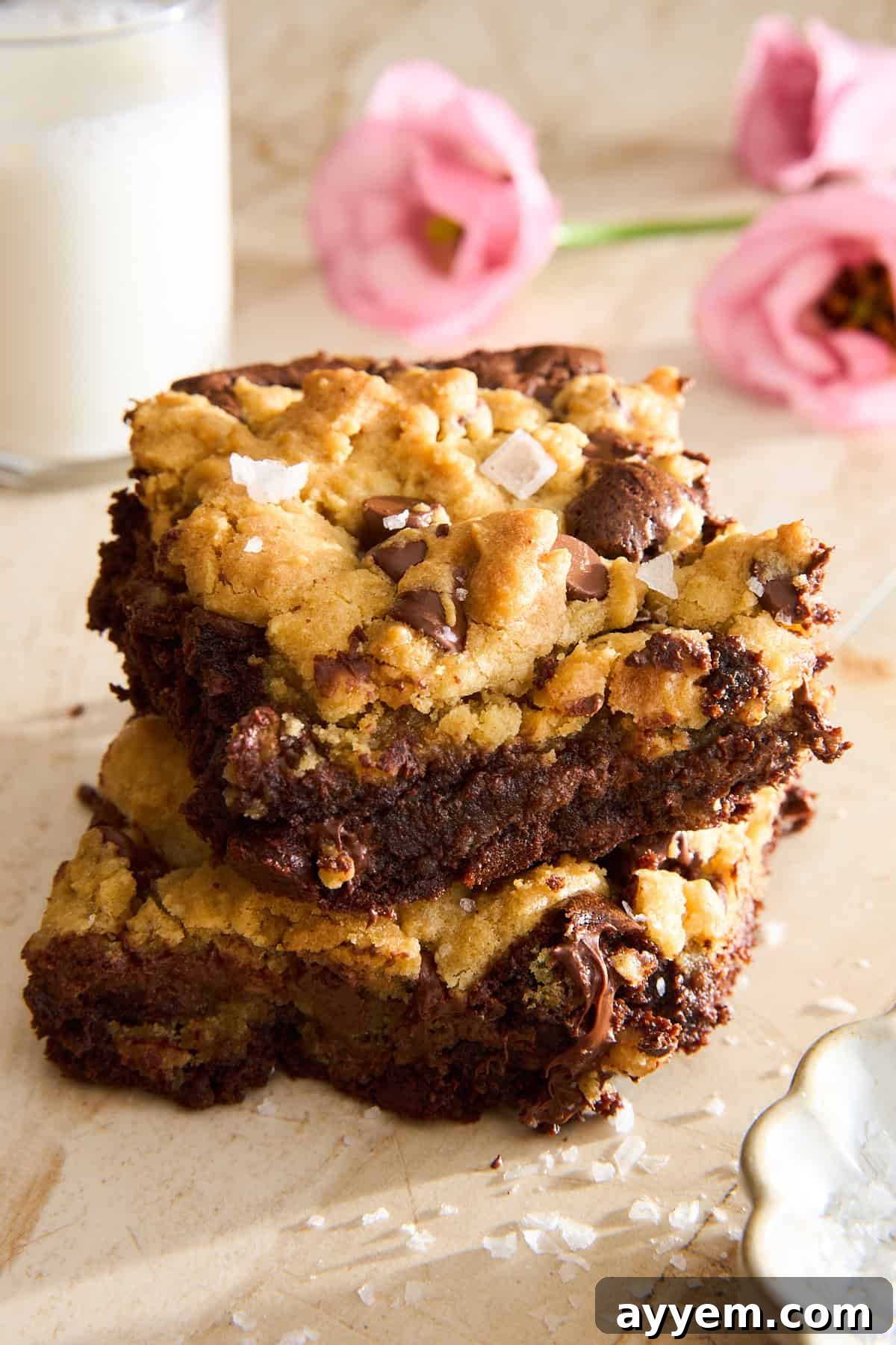 Two chocolate chip cookie bars stacked on a light surface, sprinkled with flaky sea salt. A glass of milk and pink flowers are in the blurred background.