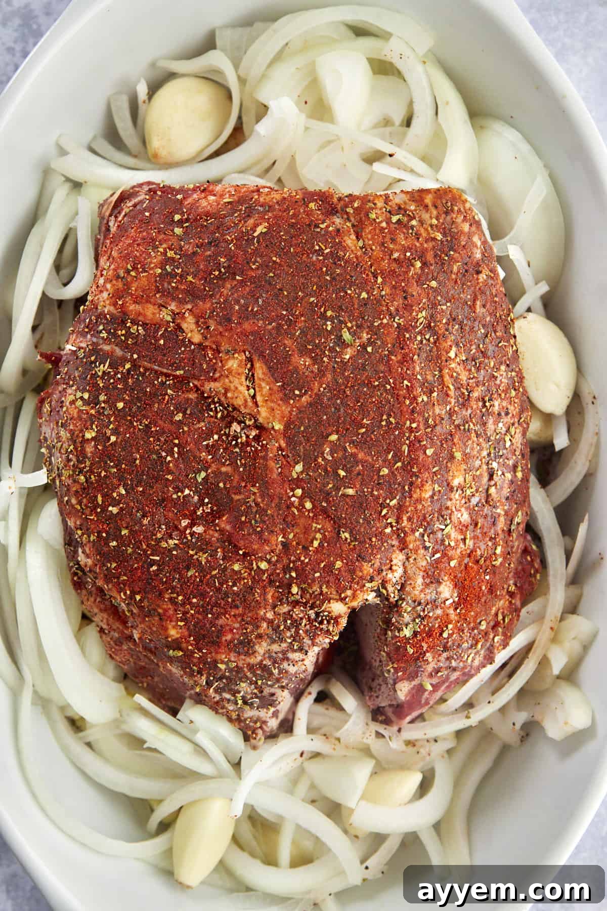 Raw chuck roast seasoned and placed on top of onions and garlic in a baking dish. 