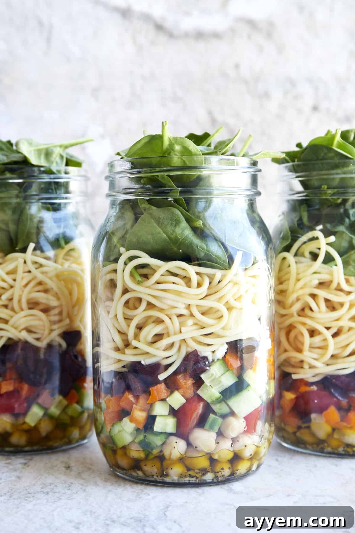 Three mason jar salads with chickpeas, veggies, spaghetti, spinach, and an olive oil dressing. They are vibrant and layered.