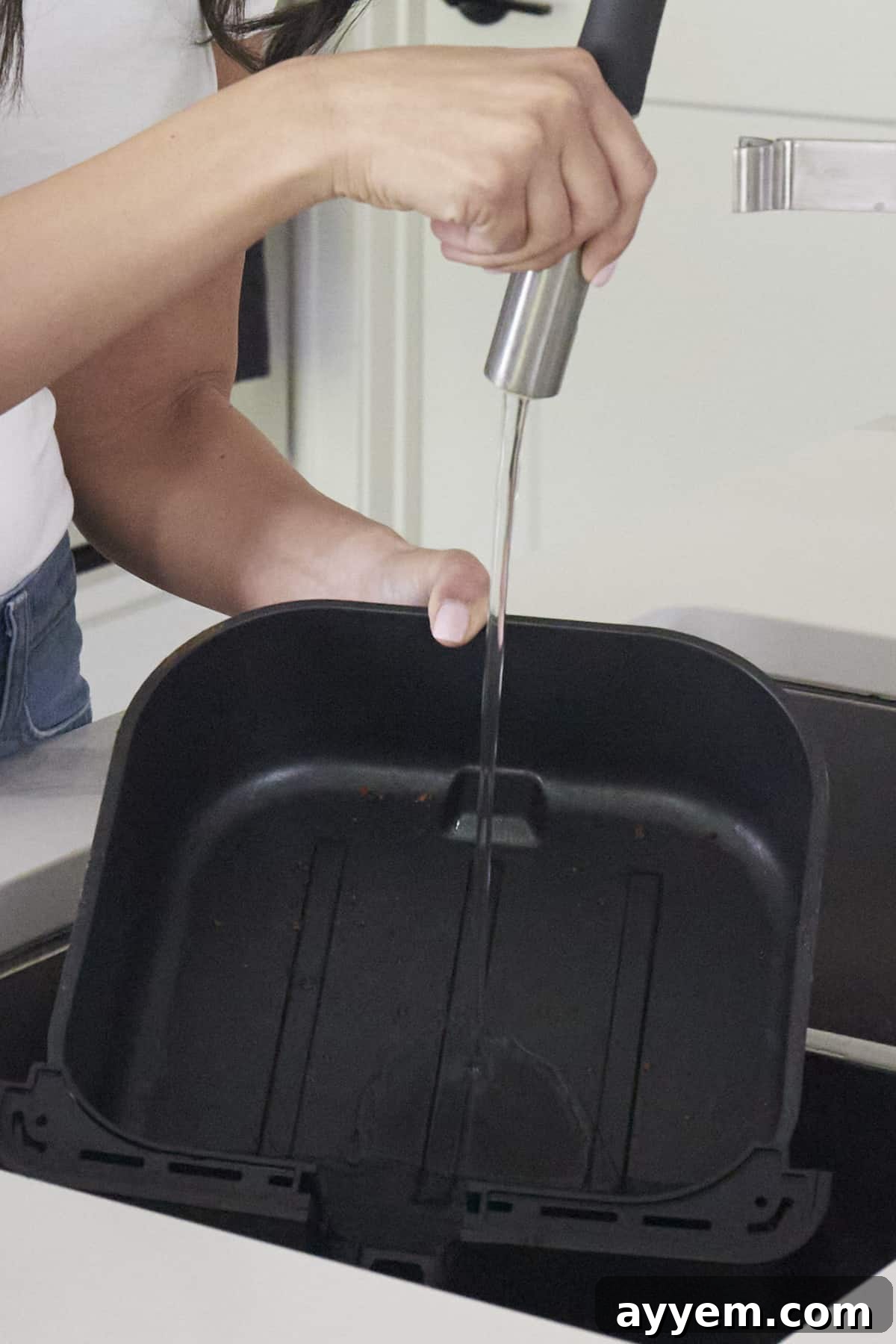 An air fryer basket being rinsed out with running water from a faucet in a sink after cleaning.
