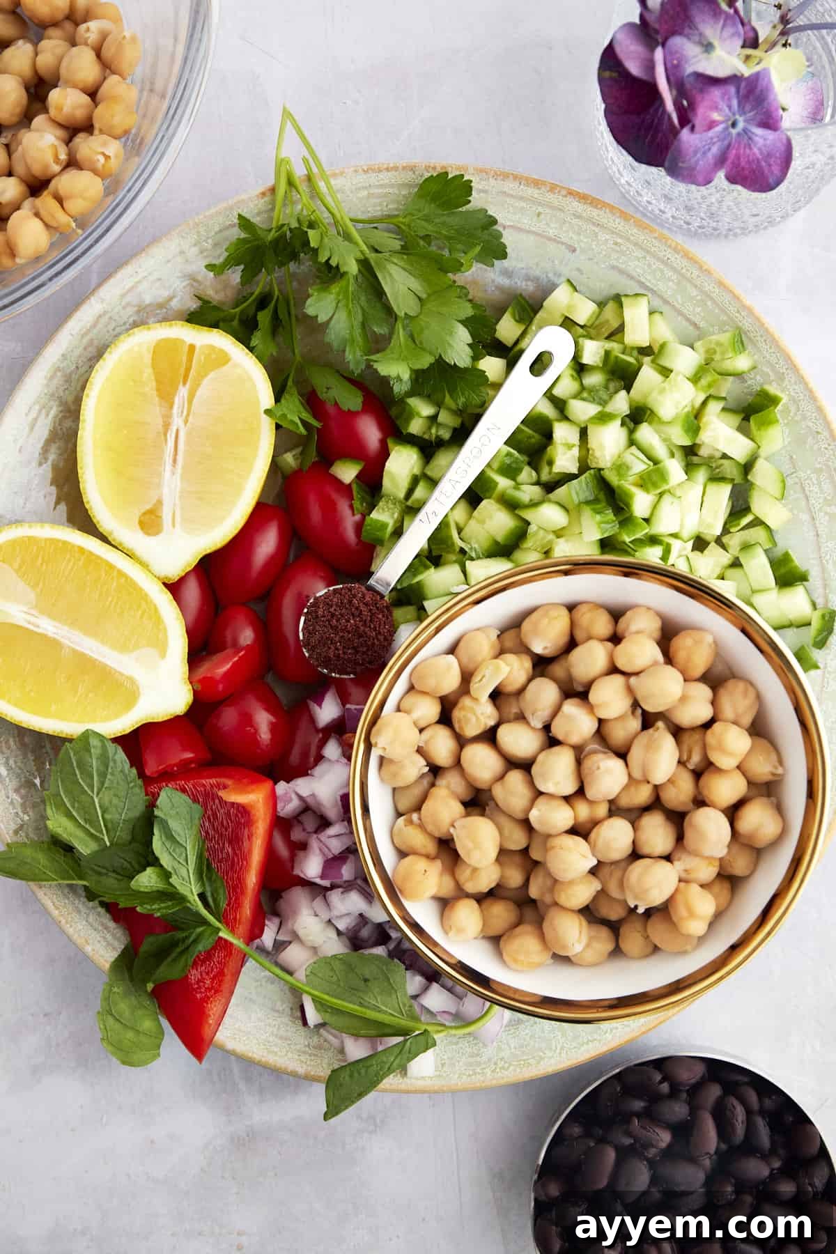 Ingredients for a Balela Salad including chickpeas, black beans, cucumbers, red onion, sumac, lemon, and fresh herbs laid out on a surface.