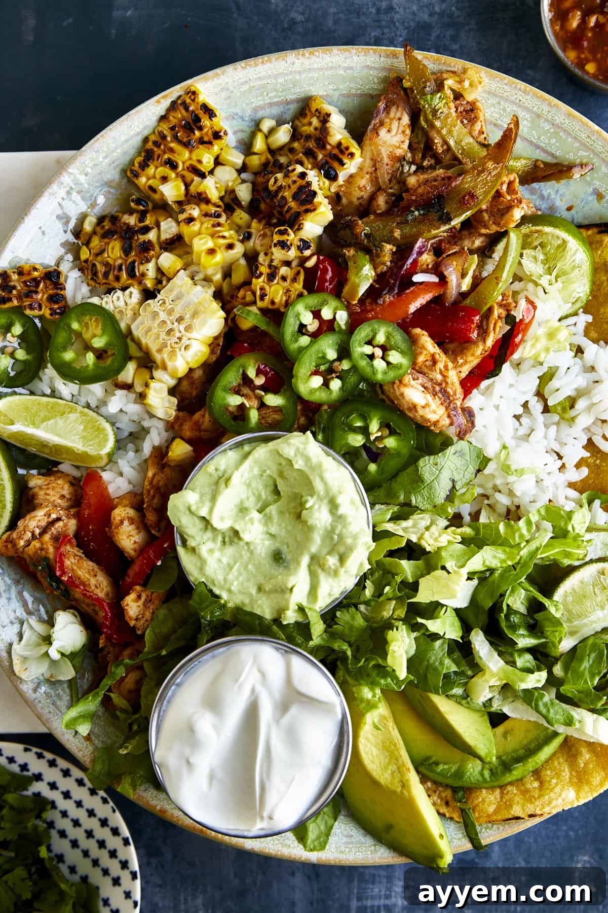 Overhead image of a chicken fajita bowl with rice, grilled corn, jalapeños, mashed avocado, sour cream, and corn tortillas. 