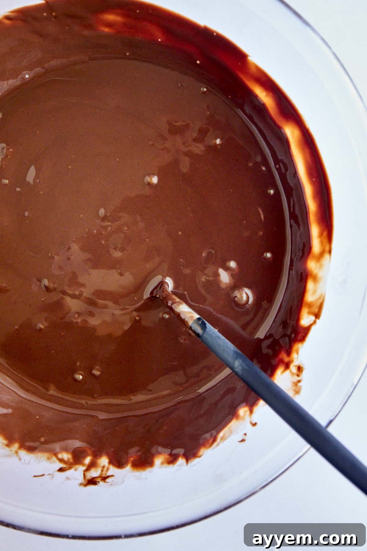 A bowl showing smooth, melted chocolate and coconut oil, with a spoon stirring the mixture.