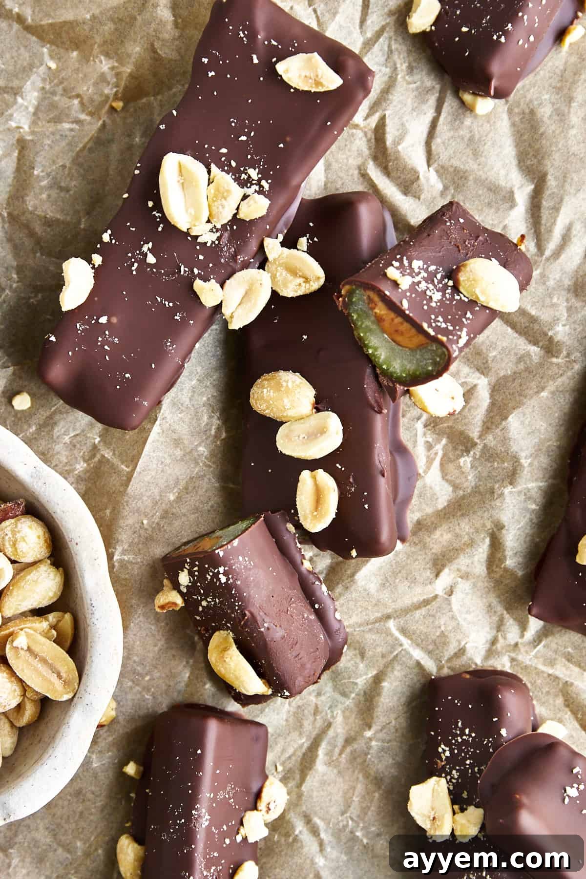 A delectable arrangement of chocolate dipped celery and peanut butter logs, beautifully topped with crushed peanuts and sea salt, resting on parchment paper. One log is artfully sliced in half, revealing the appealing layers of celery, peanut butter, and chocolate, inviting a closer look.